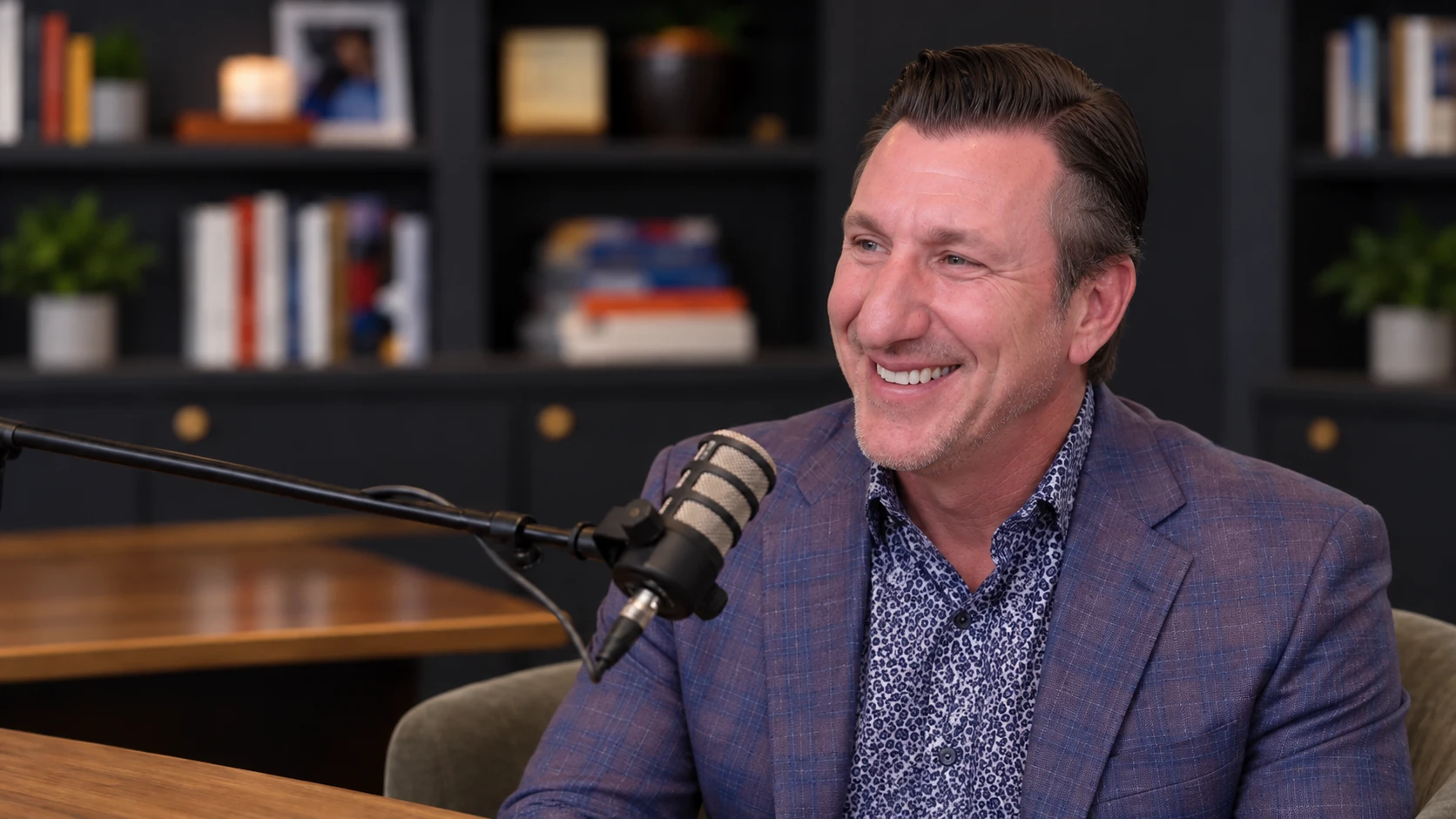 Derrick Kinney seated at a podcast microphone, with dark bookshelves behind him.